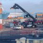 A crane lifts a container at a busy shipping yard with tractors parked nearby and a small vehicle in the foreground.